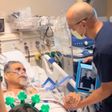 Paul Aguilar is in his hospital bed, with nurse David Yamada standing by his side, grasping his hand. Yamada is also holding a tablet with the message from Sylvester Stallone.
